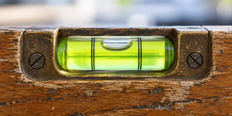 A worn wooden spirit level on a clean, pale office desk, seen so close that the glass vial and its bubble, sitting dead centre, fill the middle of the frame, with the level's body running edge to edge.