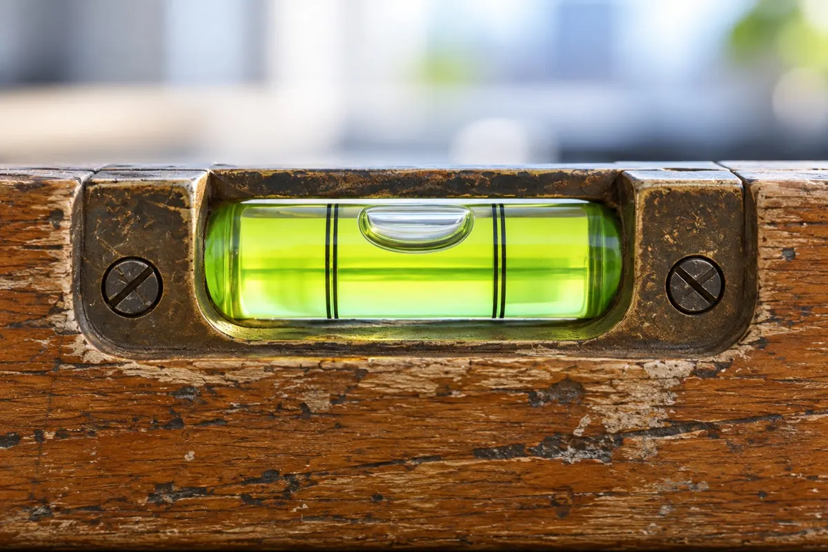 A worn wooden spirit level on a clean, pale office desk, seen so close that the glass vial and its bubble, sitting dead centre, fill the middle of the frame, with the level's body running edge to edge.